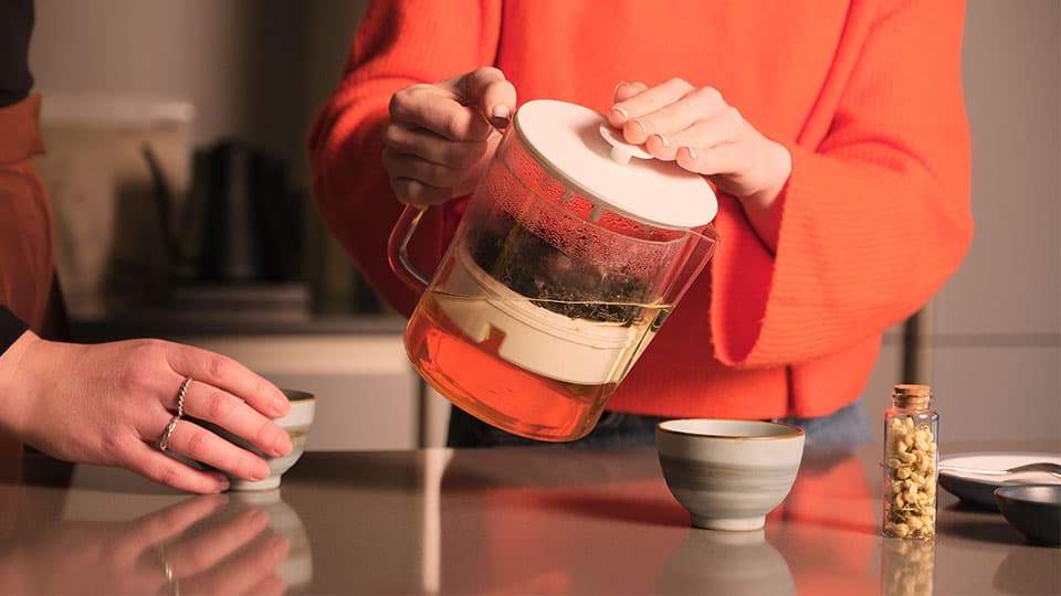 Someone wearing an orange jumper pouring from the Teapot REEDesigned. There are two grey cups on the counter and the plunger in the teapot is being pressed down and the tea leaves at the top of the water.